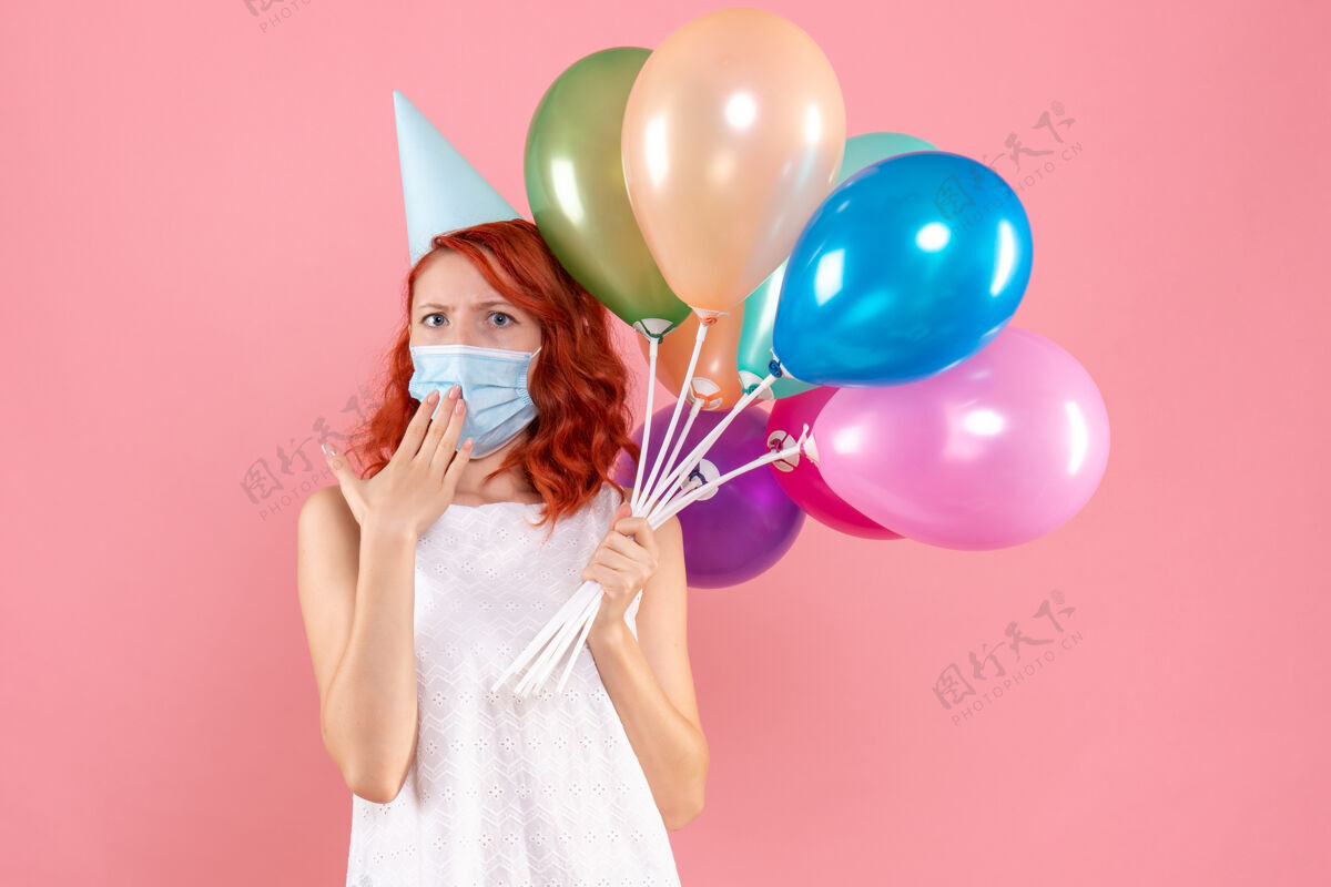粉色前视图中的年轻女子拿着五颜六色的气球在无菌面具上粉红色的墙壁女性举行Covid