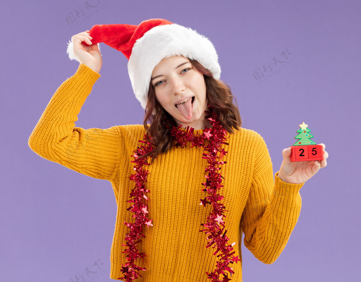 花环戴着圣诞帽 脖子上戴着花环的年轻斯拉夫女孩高兴地伸出舌头 把圣诞树装饰物孤立地放在紫色背景上 留有复制空间举行舌头Stucks