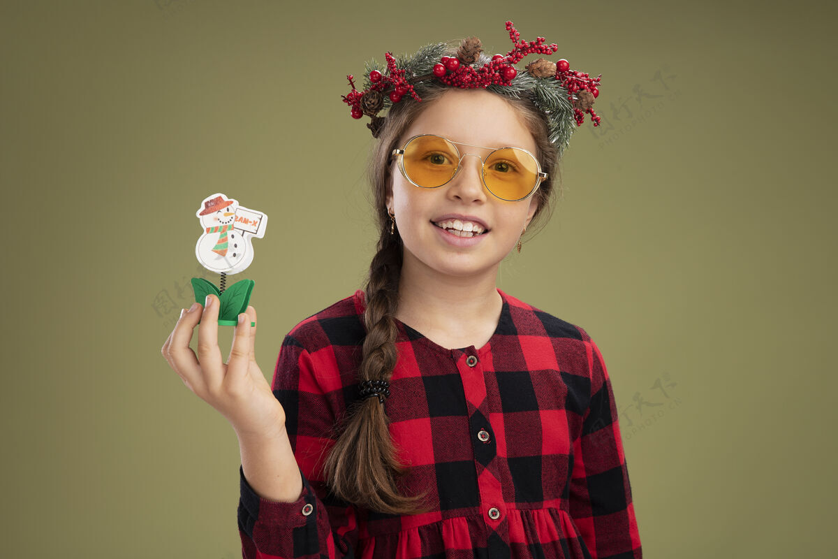 女孩小女孩戴着圣诞花圈 穿着格子裙 手里拿着圣诞玩具 站在绿色的背景下 开心而积极地看着相机积极的小微笑