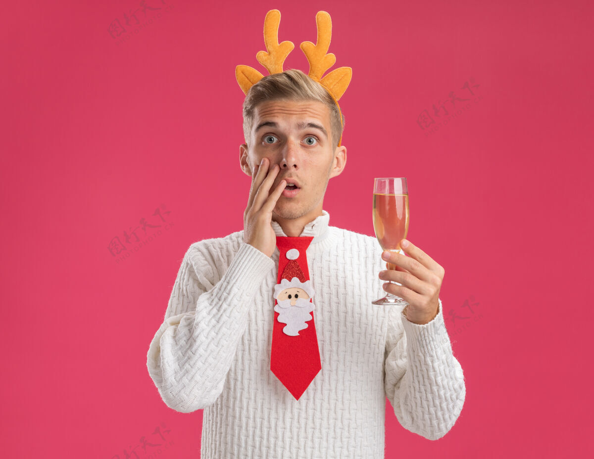 驯鹿关心的年轻帅哥戴着驯鹿鹿角头带 打着圣诞老人的领带 拿着一杯香槟 抚摸着隔离在粉色墙上的脸 留着复制空间佩戴年轻玻璃