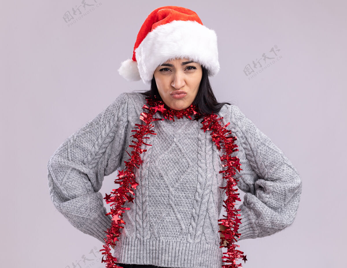 生闷气闷闷不乐的年轻白人女孩戴着圣诞帽 脖子上戴着金属丝花环 手放在腰上 看着隔离在白色背景上的摄像机女孩帽子周围
