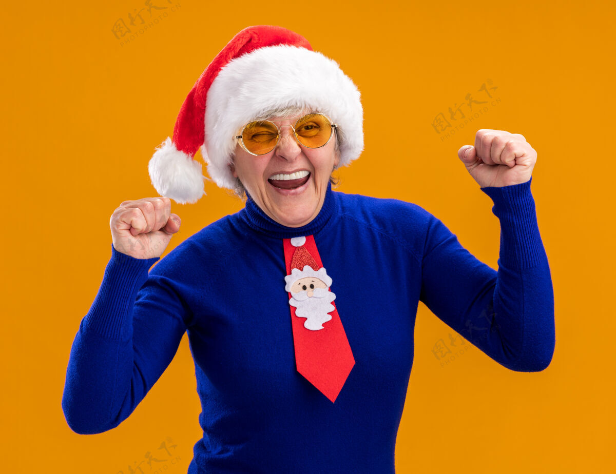 快乐戴着太阳眼镜 戴着圣诞帽 打着圣诞领带的快乐的老妇人 在橙色背景上保持拳头的隔离 并留有复制空间帽子橙色眼镜