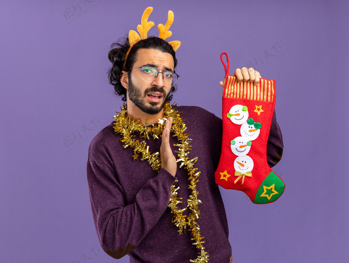 袜子年轻帅哥戴着圣诞发箍 脖子上戴着花环 手里拿着圣诞袜 手伸向蓝色背景上孤立的袜子头发手未发布