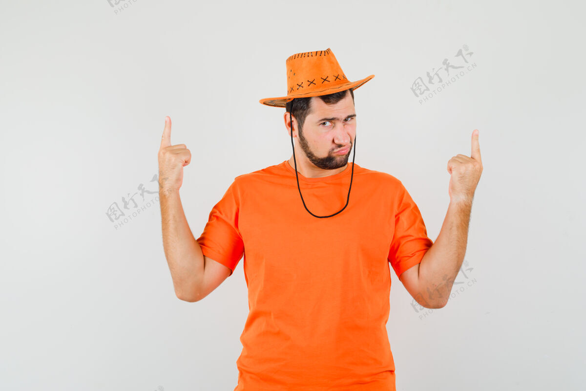 帅气年轻人指着手指在橙色的t恤衫 帽子和怀疑的样子 前视图年轻休闲衬衫