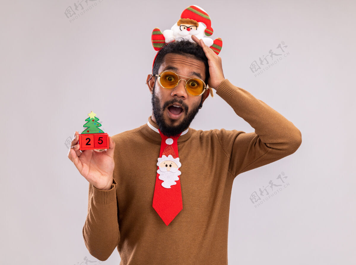 站着一个穿着棕色毛衣 头戴圣诞老人圈 打着有趣的红色领带的非洲裔美国人拿着玩具方块 上面写着日期二十五 他站在白色背景下惊讶地看着镜头非洲圣诞老人立方体