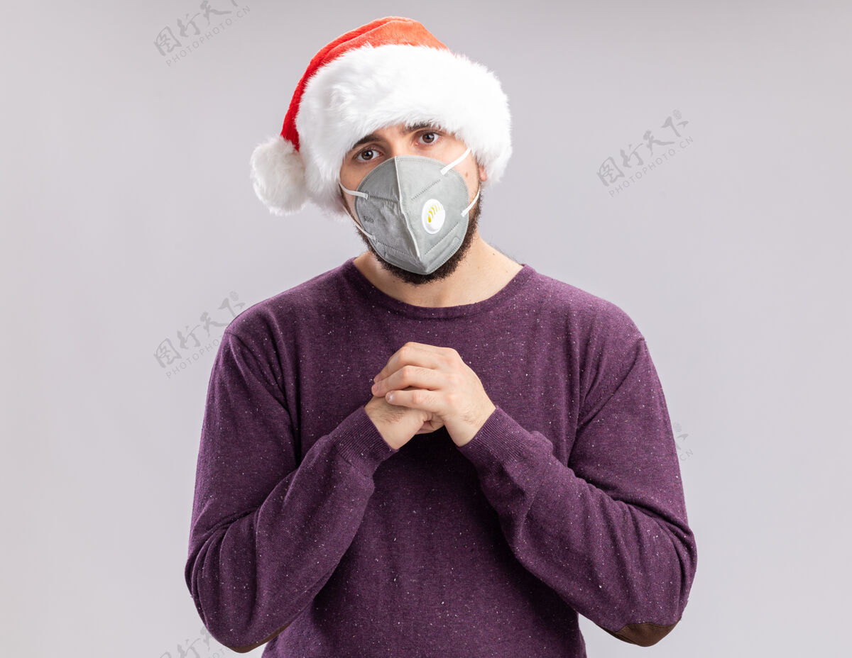 保护穿着紫色毛衣 戴着圣诞老人帽 戴着护面面具的年轻人手牵着手 站在白色背景下 看着镜头疲惫而无聊年轻站着帽子