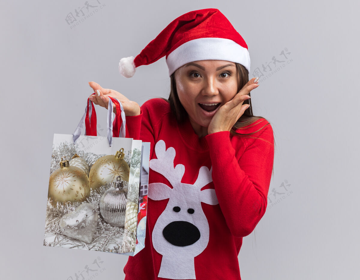 毛衣兴奋的年轻亚洲女孩戴着圣诞帽和毛衣拿着礼品袋把手放在脸颊上隔离在白色背景上兴奋包放
