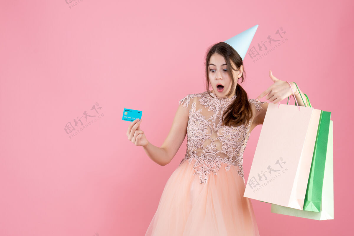 派对一个戴着派对帽的惊喜女孩 手里拿着信用卡和粉红色的购物袋信用礼服包