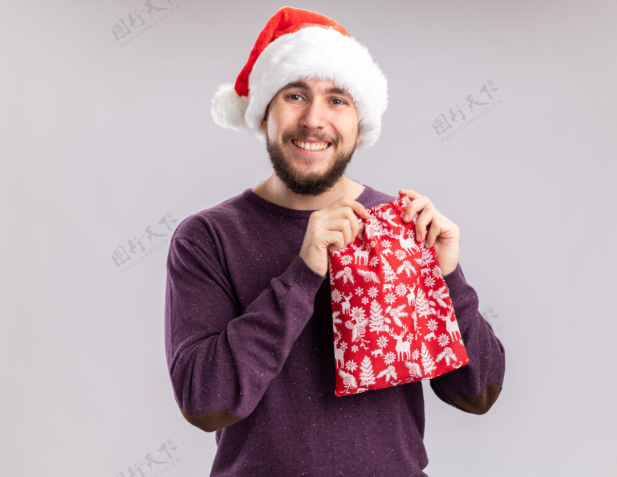 毛衣快乐的年轻人穿着紫色毛衣 戴着圣诞帽 手里拿着红包 手里拿着礼物 站在白色的背景下 看着镜头 笑容满面包宽大微笑