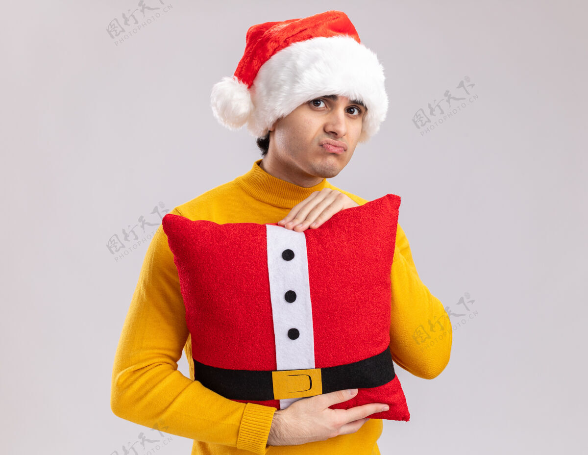 帽子一个穿着黄色高领毛衣 戴着圣诞帽 抱着圣诞枕头 站在白色背景下 愁眉苦脸地看着镜头的年轻人悲伤圣诞节枕头