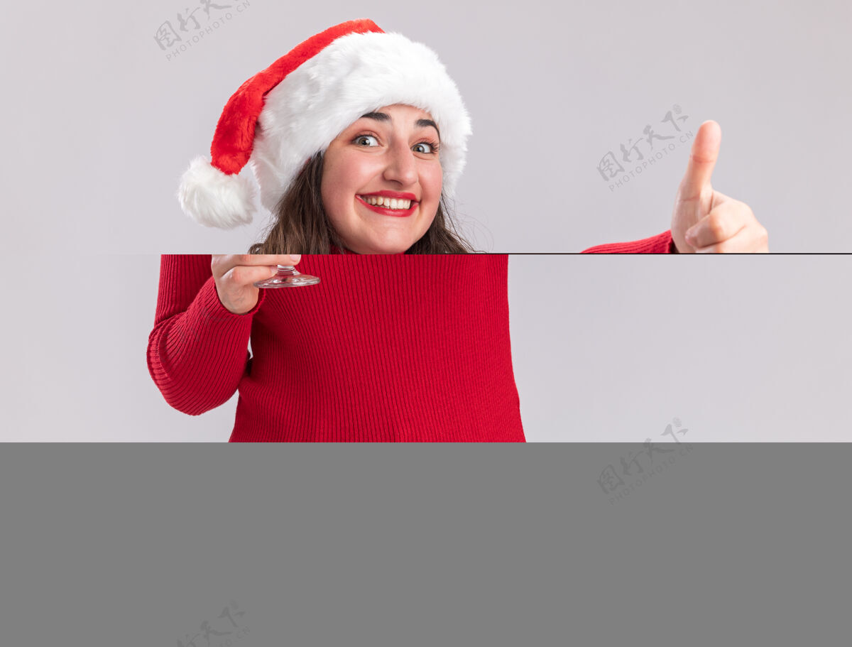 杯子快乐的年轻女孩穿着红色毛衣 戴着圣诞帽 手里拿着一杯香槟 面带微笑地看着镜头 站在白色背景上竖起大拇指年轻人站着女孩