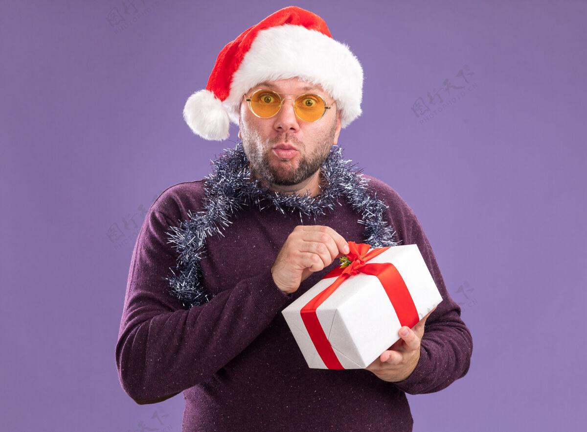 礼物好奇的中年男子戴着圣诞帽 脖子上戴着金箔花环 戴着眼镜 手里拿着礼包 手里拿着丝带 看着紫色背景上孤立的相机金属丝花环脖子