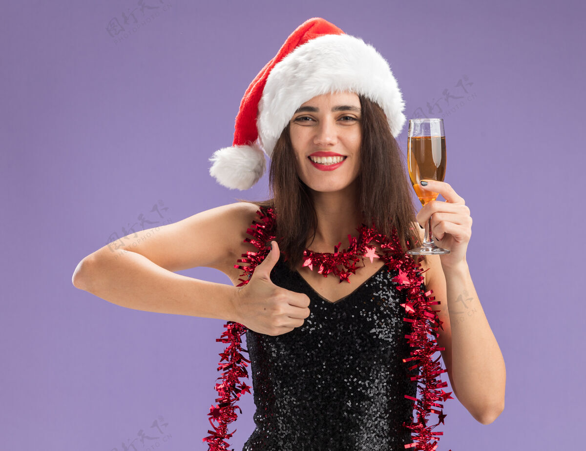 香槟微笑着的年轻漂亮女孩戴着圣诞帽 脖子上戴着花环 手里拿着一杯香槟 在紫色的背景上孤立地竖起大拇指微笑脖子花环