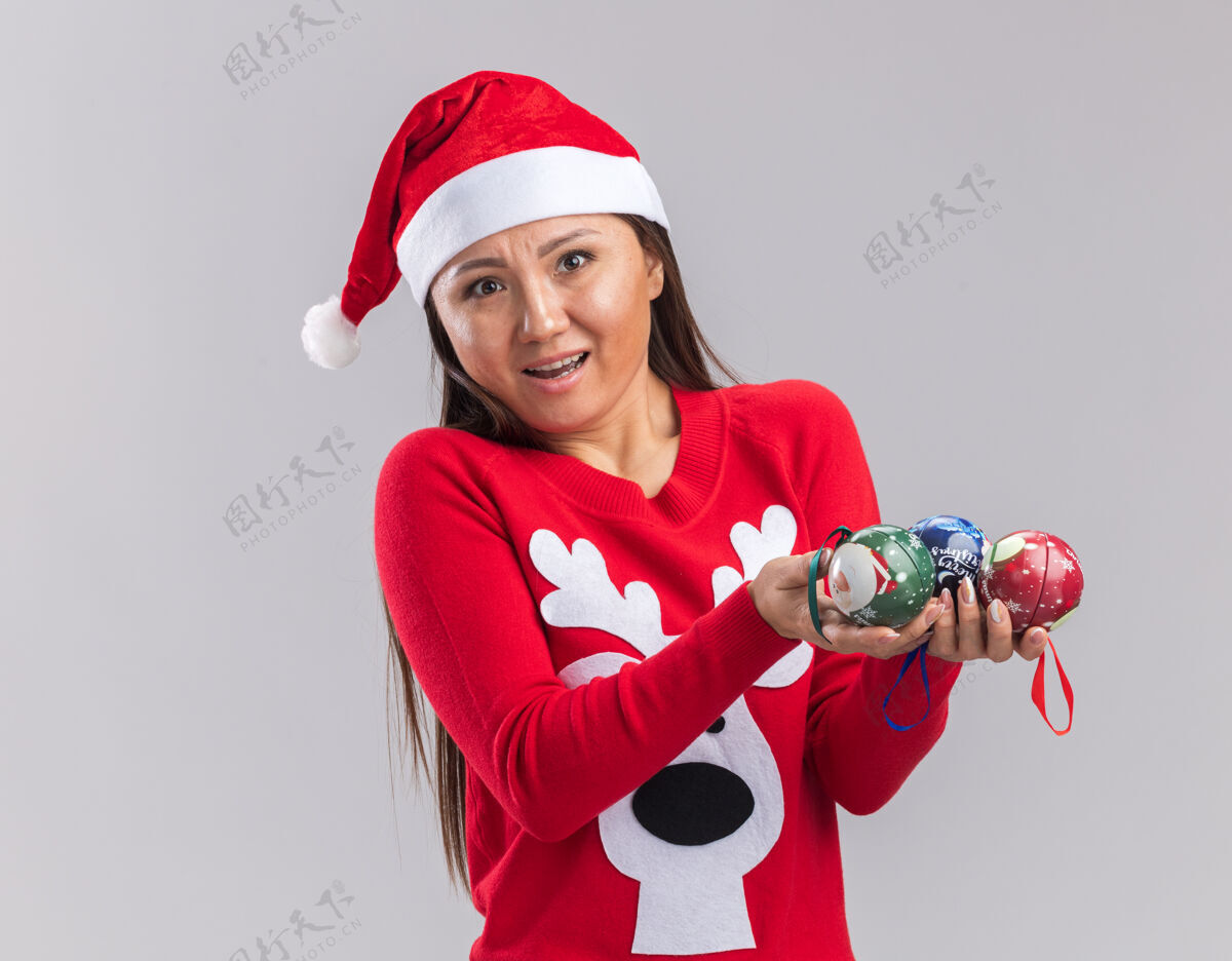 亚洲人惊讶的年轻亚洲女孩戴着圣诞帽和毛衣拿着圣诞树球孤立的白色背景帽子毛衣年轻