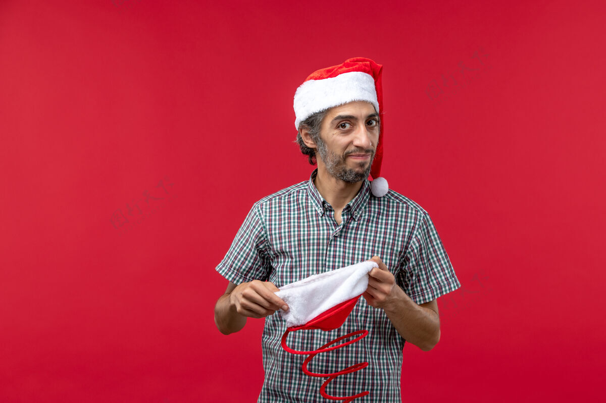 圣诞节红墙上戴着圣诞玩具帽的年轻人的正视图肖像帅哥前面