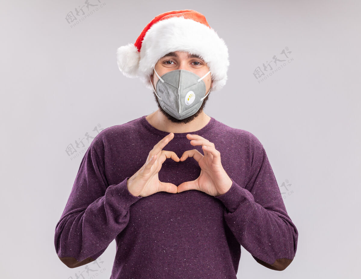 制作一个穿着紫色毛衣 戴着圣诞帽 戴着护面面具的年轻人看着镜头 用手指在白色背景上做着心形手势毛衣帽子手指