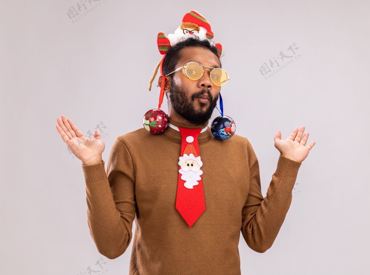 轮辋一个非洲裔美国人 头上戴着圣诞老人的戒指 头上系着一条有趣的红色领带 耳朵上戴着圣诞球 看起来很困惑 他举起双臂站在白色的背景上美国站着圣诞节