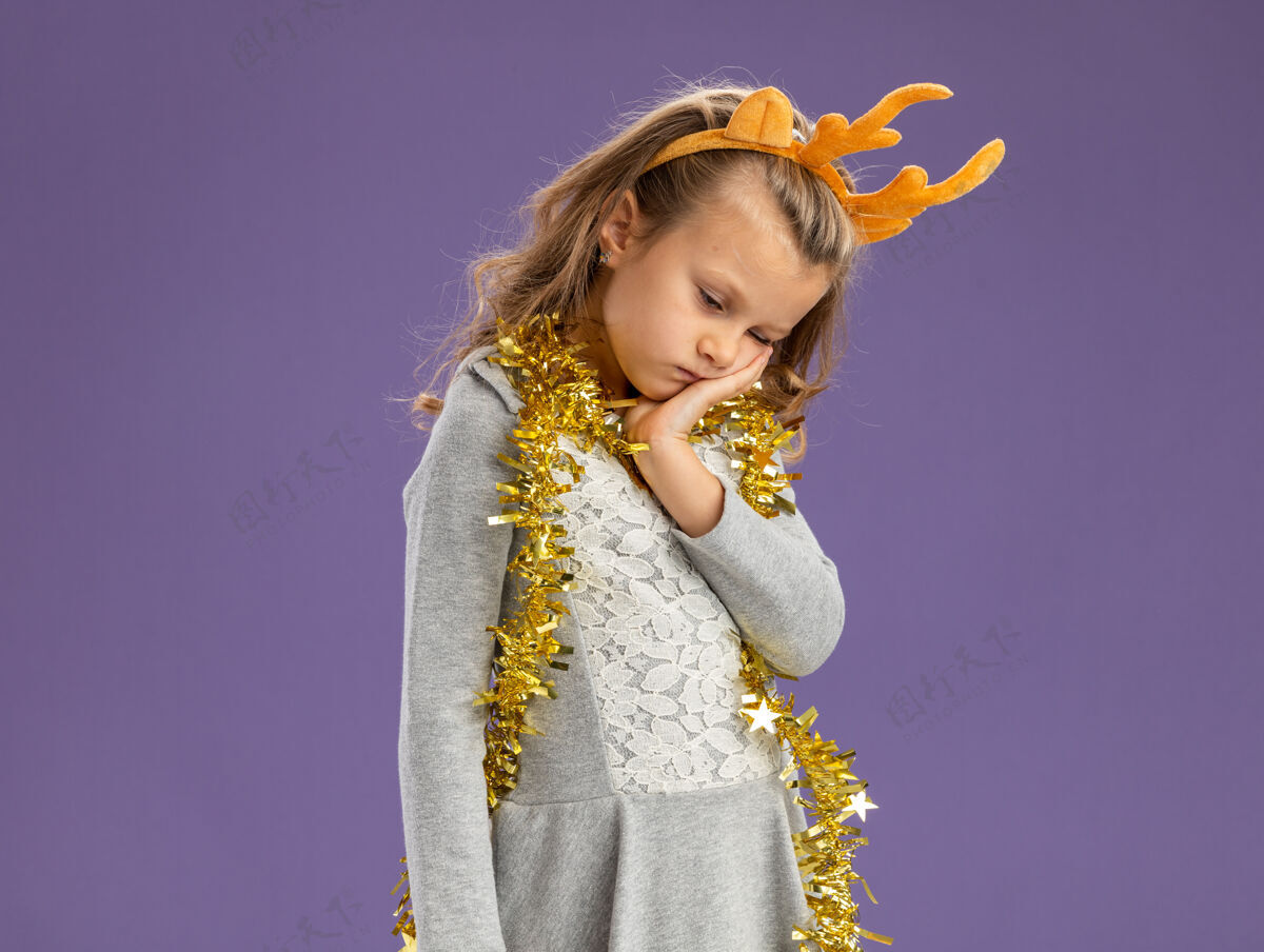 悲伤悲伤的低着头小女孩戴着圣诞发箍 脖子上戴着花环 手放在脸颊上 与蓝色背景隔离 留有复制空间脖子手花环