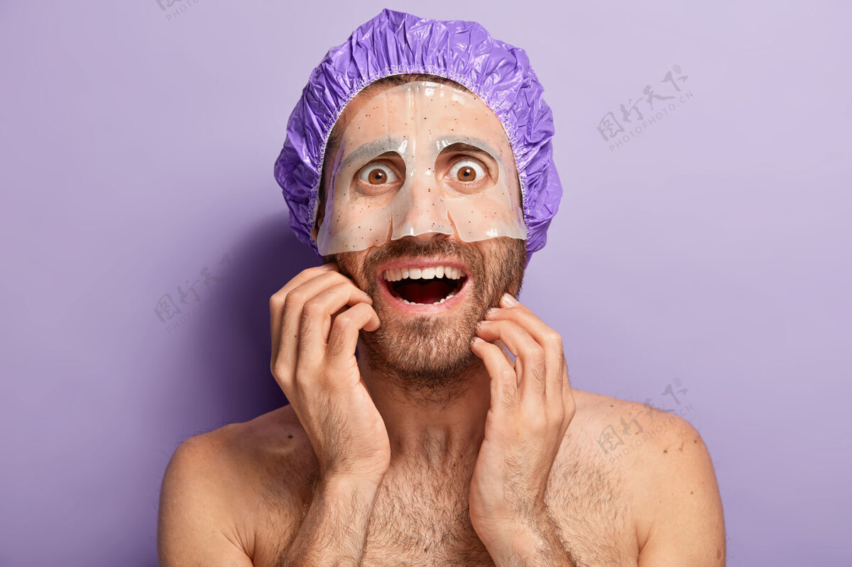 凝视裸体快乐男人在脸上涂保湿面膜 戴紫色淋浴帽 享受美容护肤面部护理 有棕色的眼睛 光着肩膀躯干男性肌肉
