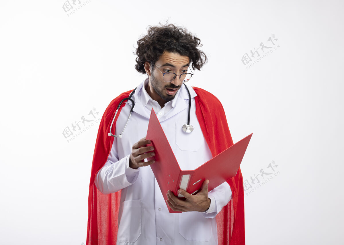 脖子惊讶的年轻白种人超级英雄戴着眼镜 身穿医生制服 披着红色斗篷 脖子上戴着听诊器 拿着并看着文件夹和复印空间惊讶文件夹医生