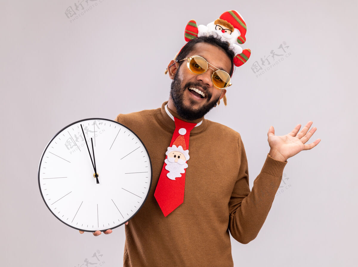 领带非洲裔美国人 身穿棕色毛衣 头戴圣诞老人戒指 打着有趣的红色领带 拿着时钟看着摄像机 兴高采烈地微笑着 举起手臂站在白色背景上微笑轮辋胳膊