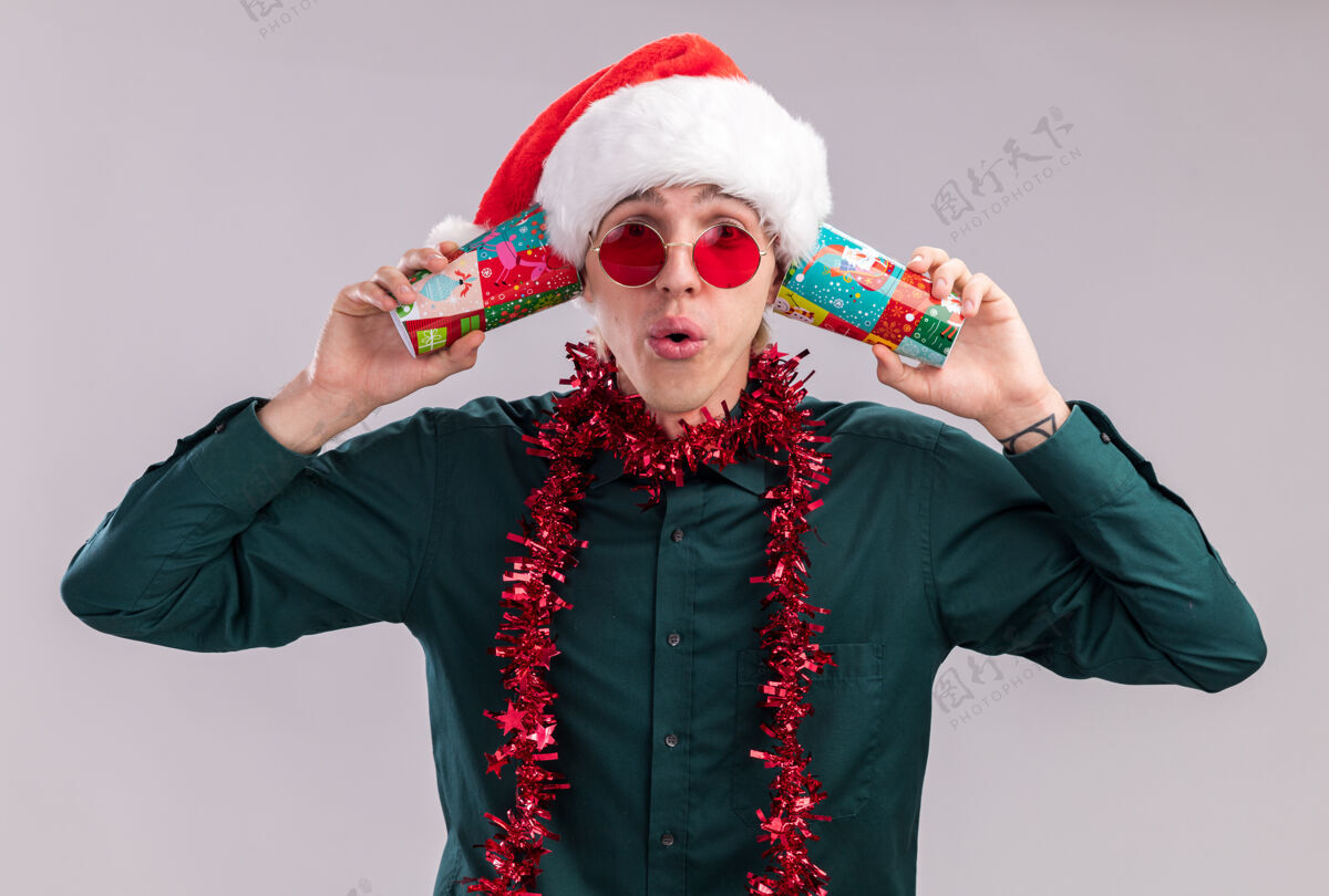 圣诞节令人印象深刻的年轻金发男子戴着圣诞帽 戴着眼镜 脖子上戴着金属丝花环 耳边拿着塑料圣诞杯 听着秘密 看着隔离在白色背景上的相机塑料花环圣诞老人