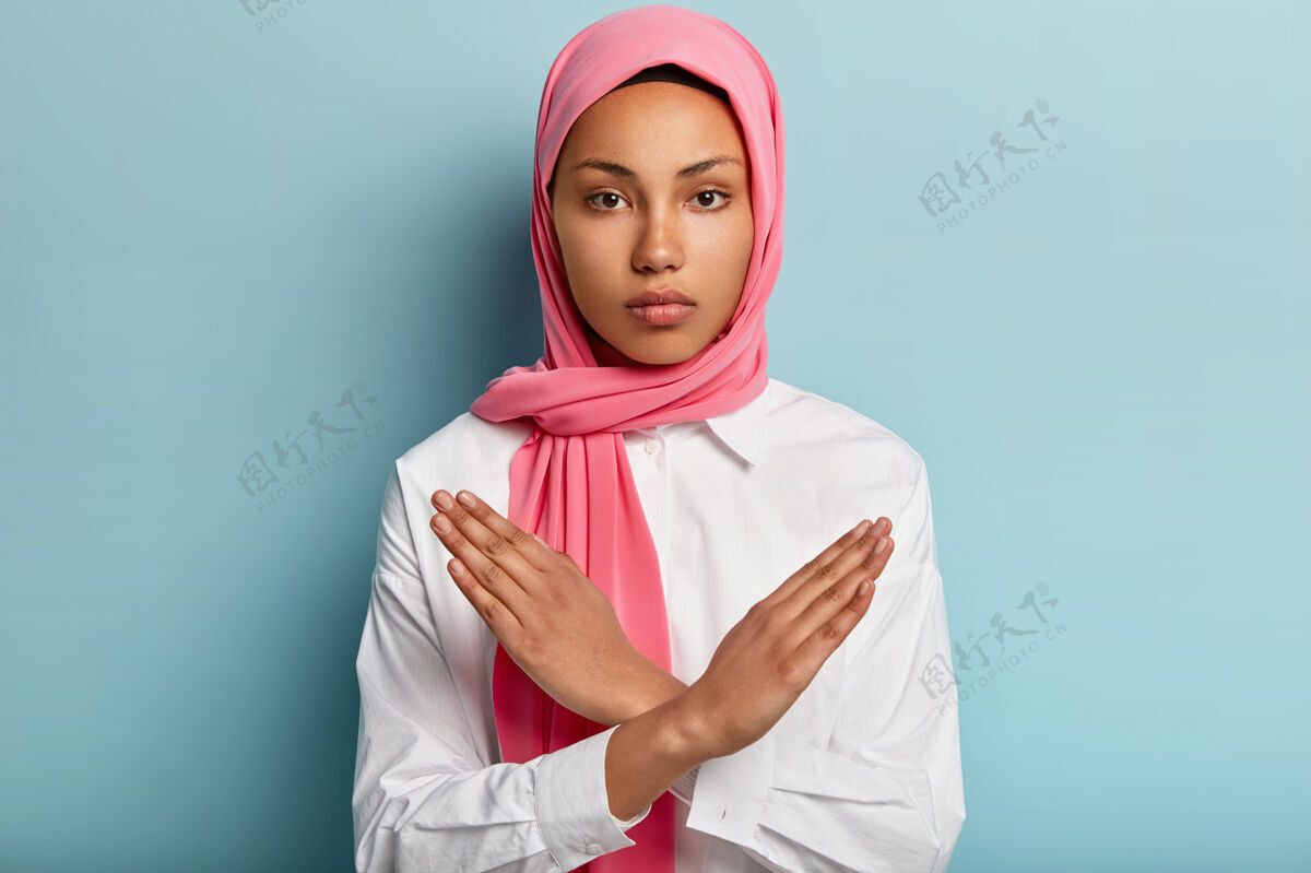 玫瑰色严肃穆斯林妇女的室内镜头做出否认手势 双臂交叉放在胸前 展示停车标志 戴头巾 遵循宗教着装规范 隔离在蓝色墙壁上宗教阿拉伯语否认