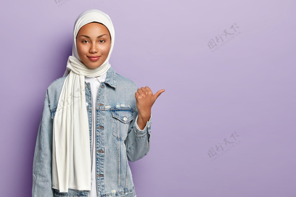 牛仔身着传统阿拉伯服装的迷人女性 拇指指向右侧 在空白处呈现物体 有宗教观点 孤立于紫色墙壁之上宗教概念穆斯林摆姿势宗教