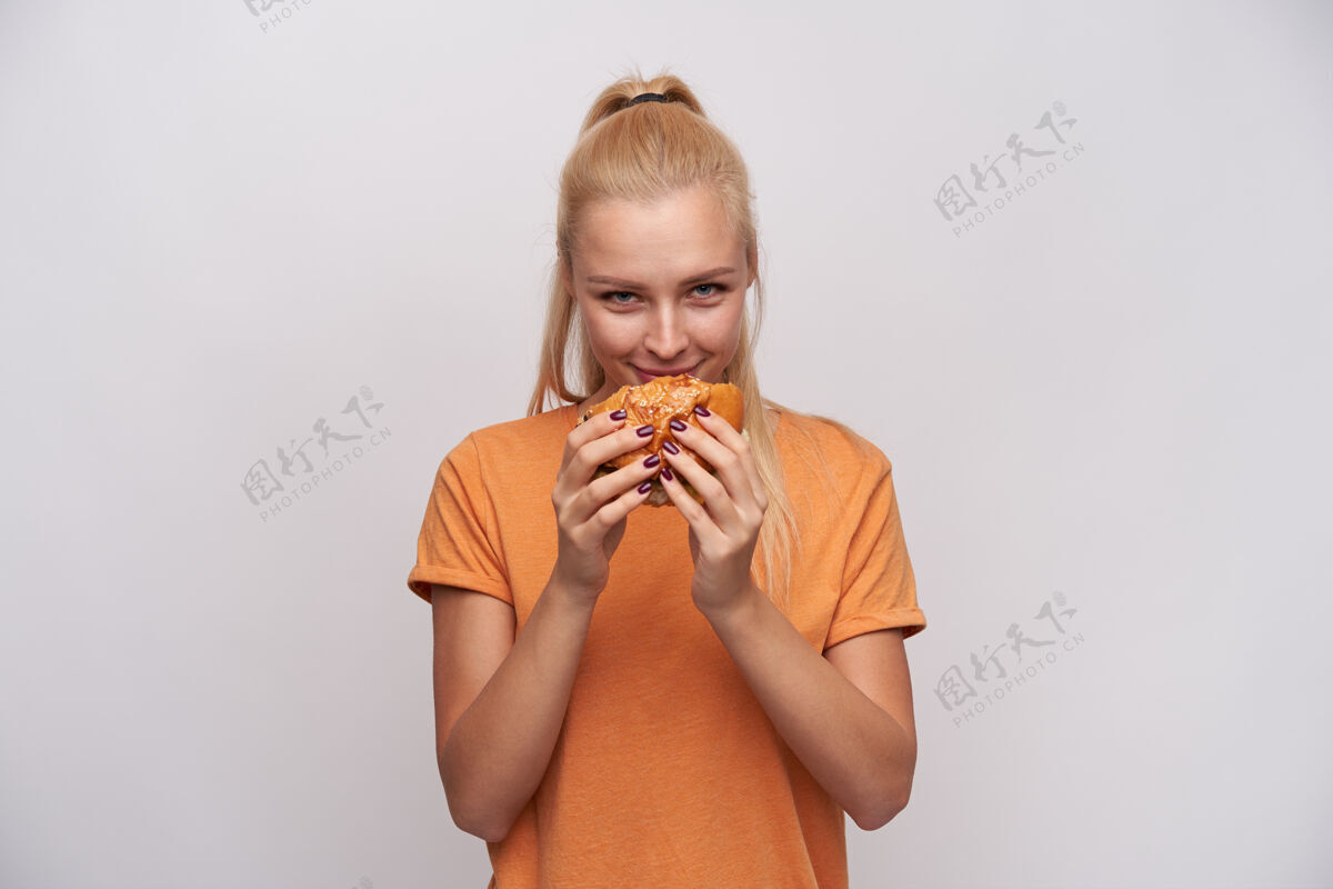 休闲满意的年轻迷人的金发女性 扎着马尾辫 吃着新鲜的汉堡 兴高采烈地看着镜头 穿着橙色t恤 站在白色背景下摆姿势垃圾美甲
