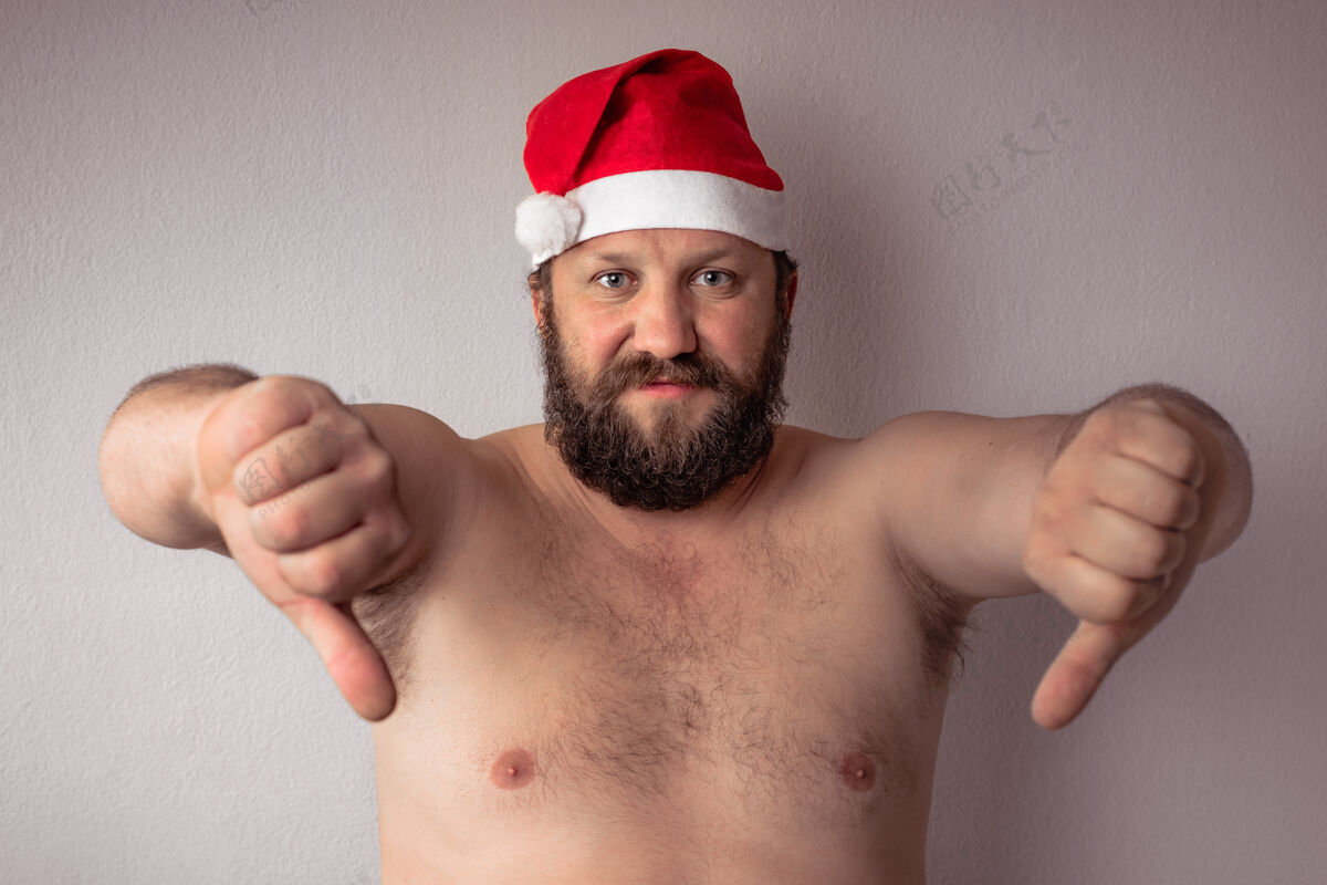 向下满脸胡须的半裸男人戴着圣诞老人的帽子 大拇指朝下靠在灰色的墙上成人帽子庆祝