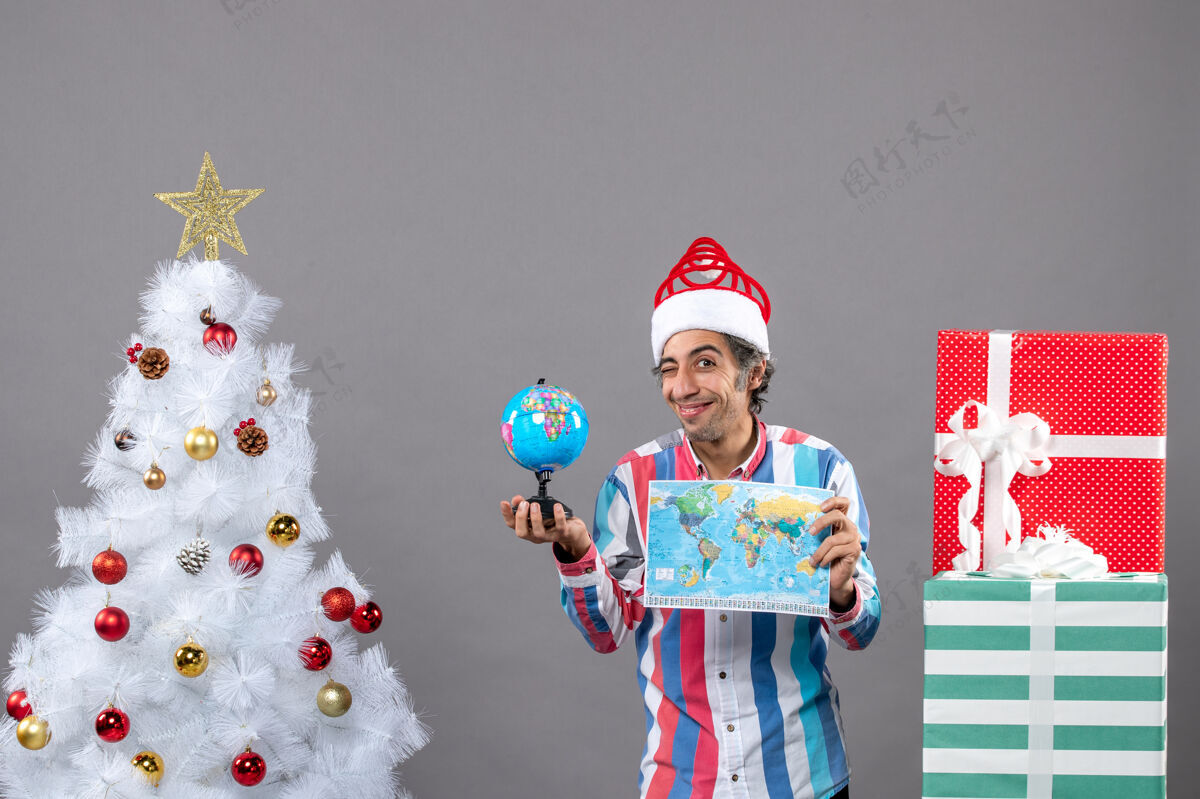 庆祝正面图微笑的男人眨着眼睛拿着世界地图和地球仪圣诞树男人旅行者