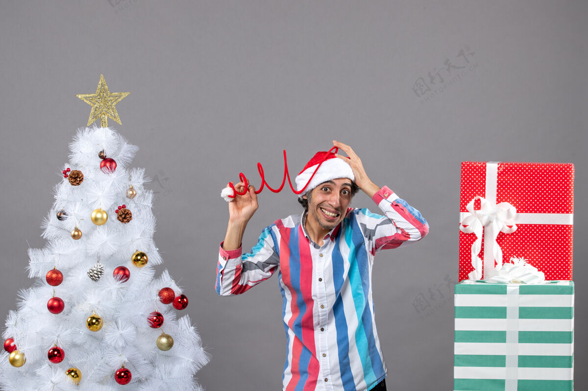 快乐前视图惊讶的年轻人拿着他的螺旋弹簧圣诞帽靠近白色圣诞树人人微笑