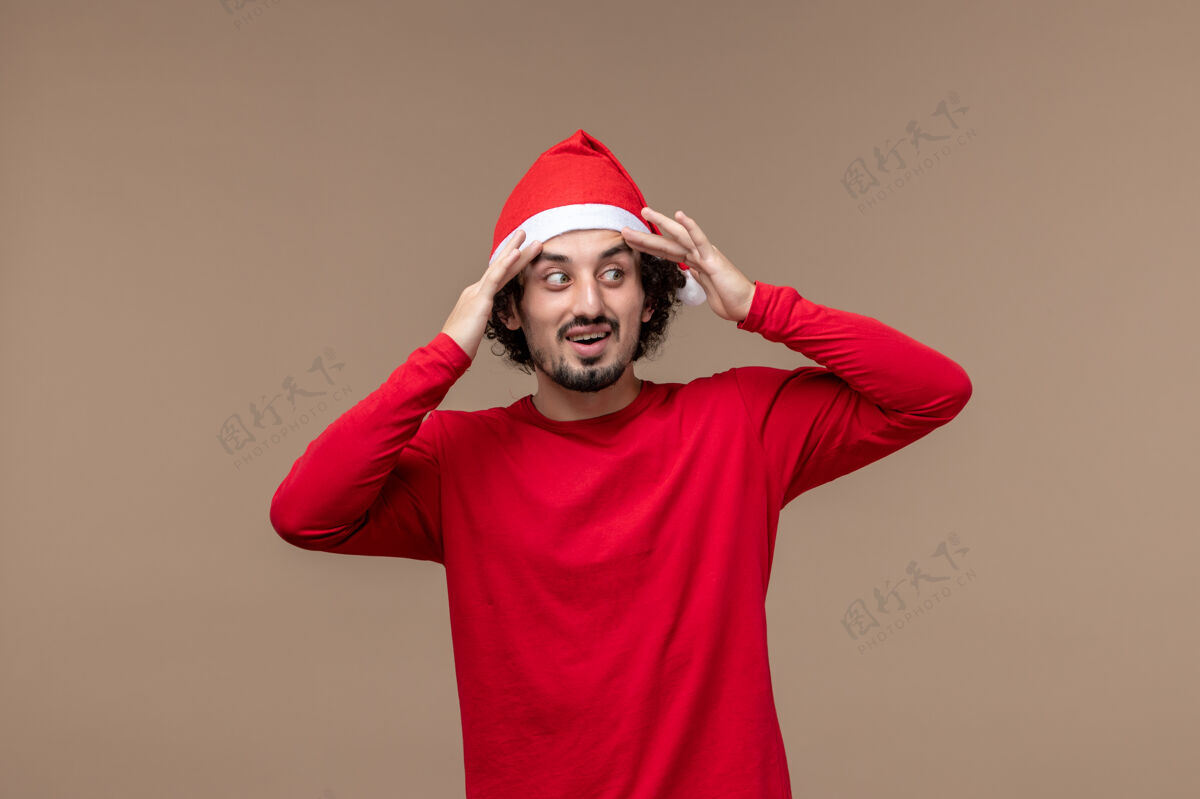 节日正面图棕色背景上穿着红色斗篷的年轻男性感慨圣诞假期帽子前面微笑