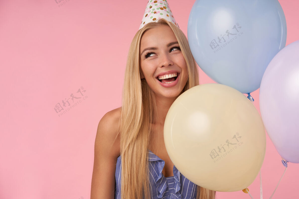 休闲特写镜头中迷人的年轻金发女性一边看一边 面带微笑 在蓝色夏装和生日帽的五彩缤纷的气球里摆姿势 孤立在粉色背景下庆祝裙子气球