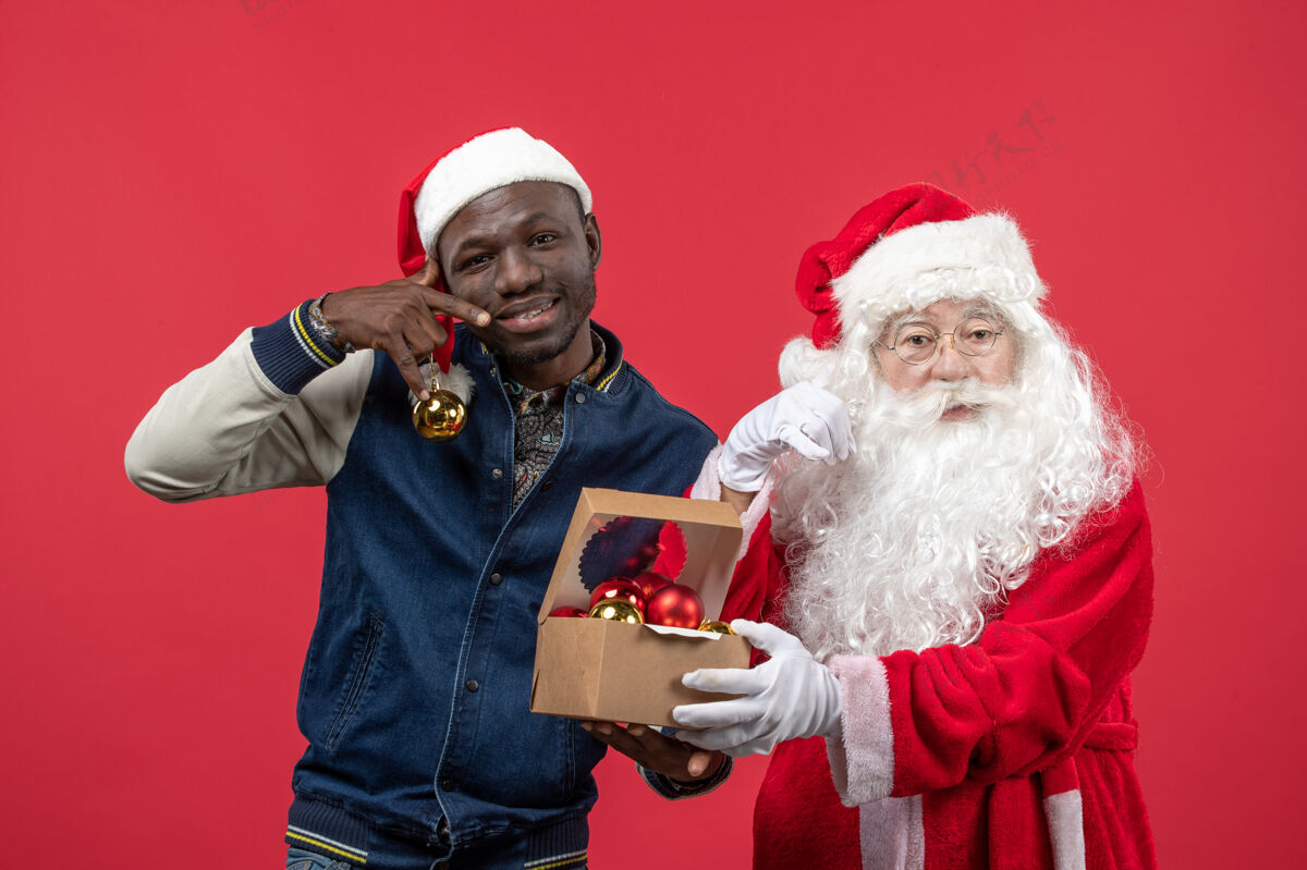 快乐红墙上的圣诞老人和拿圣诞树玩具的年轻人的正视图节日男人服装