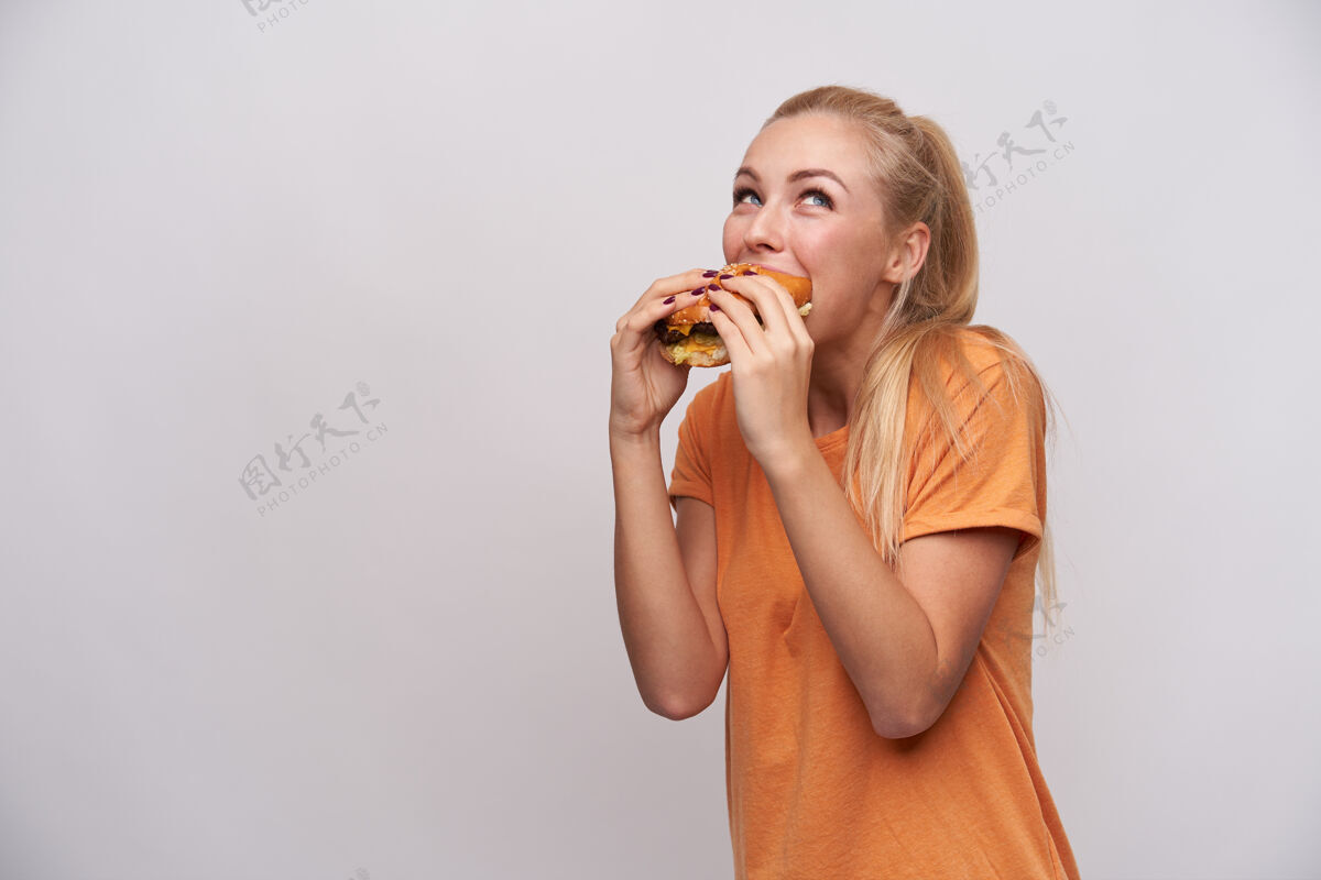 T恤一张室内照片 一位年轻开朗的金发女郎 留着随意的发型 一边在白色背景下穿着橙色t恤摆姿势 一边愉快地吃着汉堡包穿着20多岁美甲