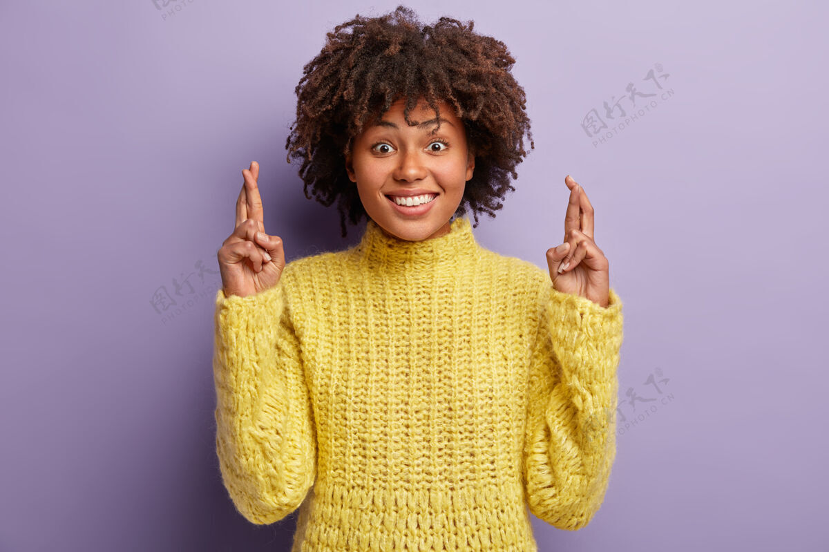 祈祷超级黑皮肤的女性交叉手指 笑容灿烂 祈求美好的事物 希望她的梦想成真 穿着暖黄色的套头衫 在紫色的墙壁上摆姿势愿望的概念愿望希望幸福