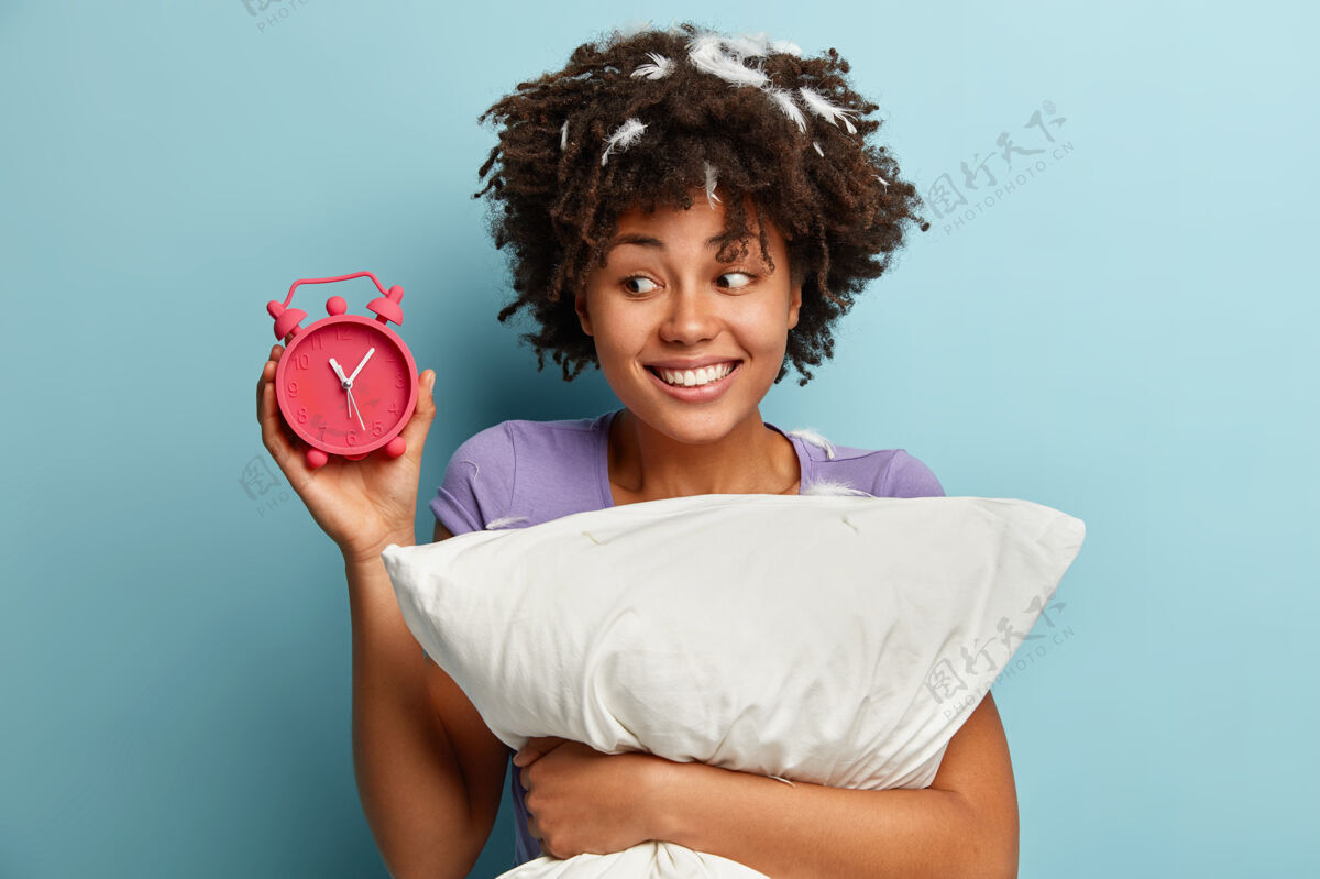 年轻可爱的非洲裔美国女人的照片 卷发 闹钟上显示时间 很高兴晚上睡得很长 微笑积极 抱着柔软的白色枕头 隔着蓝色的墙壁睡觉睡眠开放风格