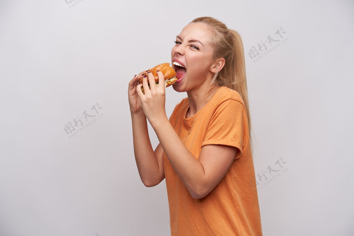情绪摄影棚拍摄了一位年轻漂亮的金发女郎 一头随意的发型 手里拿着汉堡包 正准备从汉堡包上咬下一块 张大嘴巴贪婪地看着镜头微笑T恤天然