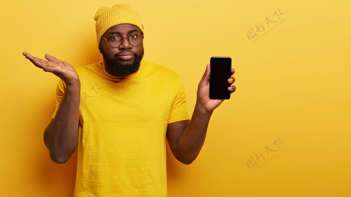 设备犹豫迷茫的男人选择新的智能手机 拿着模拟屏幕的现代电子设备 疑惑地举起手掌 犹豫是否购买 戴着鲜艳时尚的黄色帽子和t恤非洲犹豫困惑