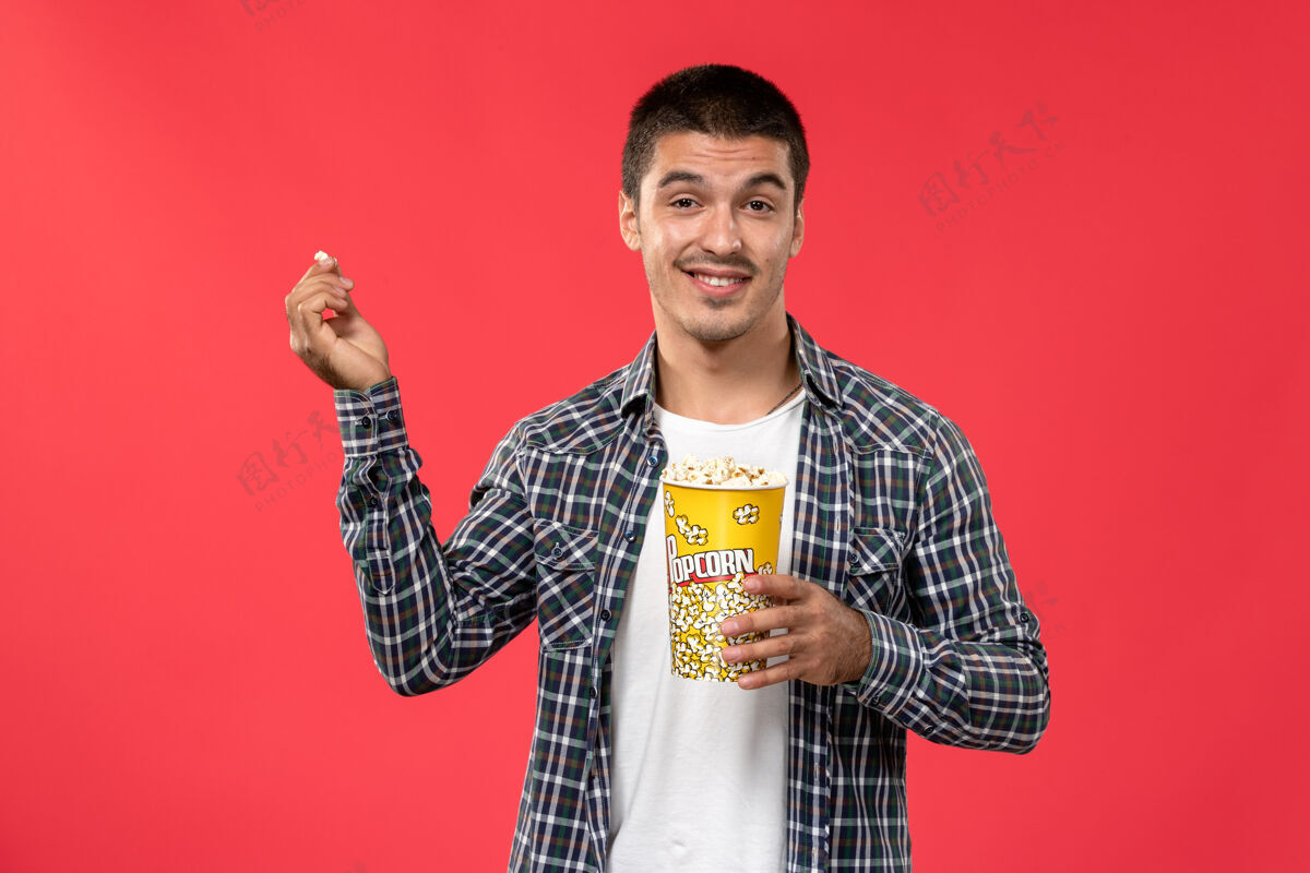 男性正面图年轻男子拿着爆米花包 脸上带着喜悦的表情在淡红色的墙上看男性电影表演者电影电影