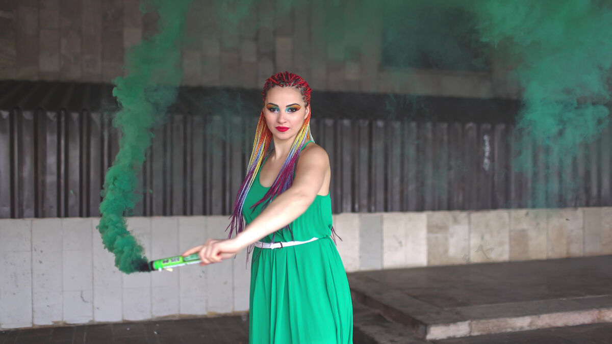 化妆一个年轻开朗的女孩 穿着亮绿色的春装 带着亮片和彩虹色的非洲辫子她站在一栋建筑附近 在街上吹着绿色的烟多彩漂亮姿势