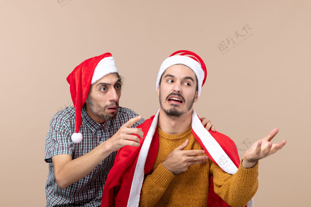 圣诞老人前视图两个戴着圣诞帽的感兴趣的家伙在看米色孤立背景上的东西微笑微笑看