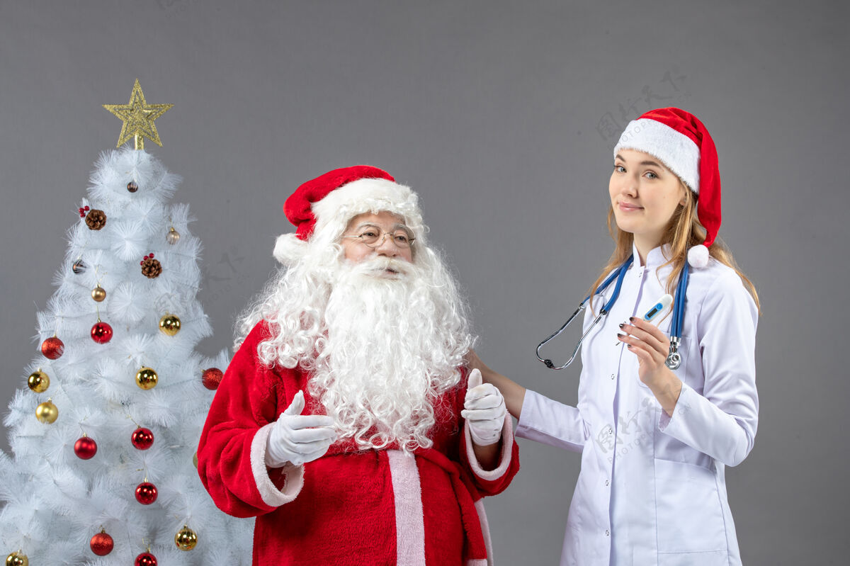 节日圣诞老人和女医生的前视图 女医生拿着测温仪在灰色的墙上圣诞老人女性圣诞节