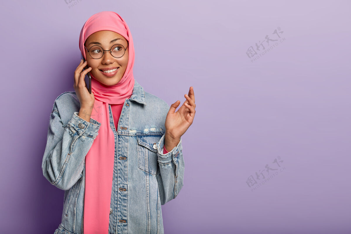 微笑正面深色皮肤的女性头上戴着传统围巾 把手机放在耳边 享受愉快的交谈 一边做手势 一边向孤立在紫色地面上的对话者讲述一些激动人心的事情伊斯兰满意手机
