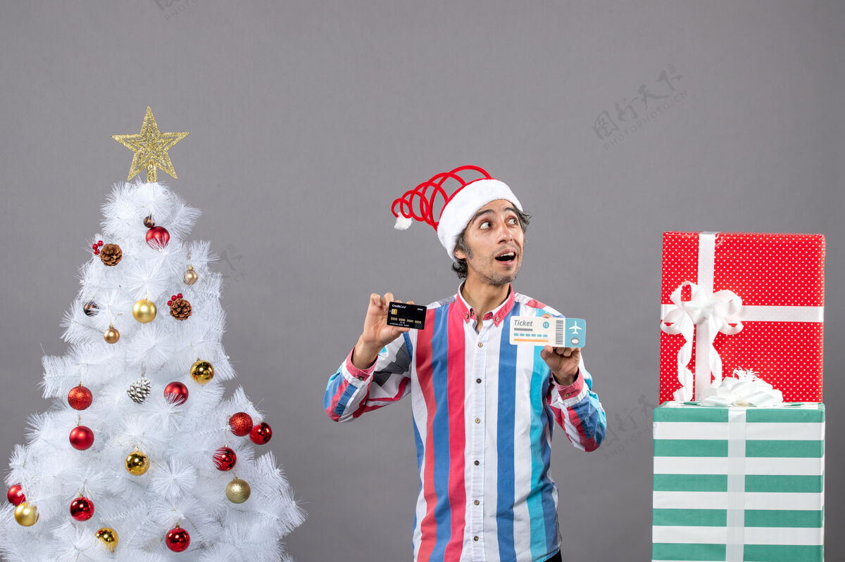 快乐前视图：一个眼睛敏锐的男人拿着卡片和旅行票 围着圣诞树和礼物圣诞周围树