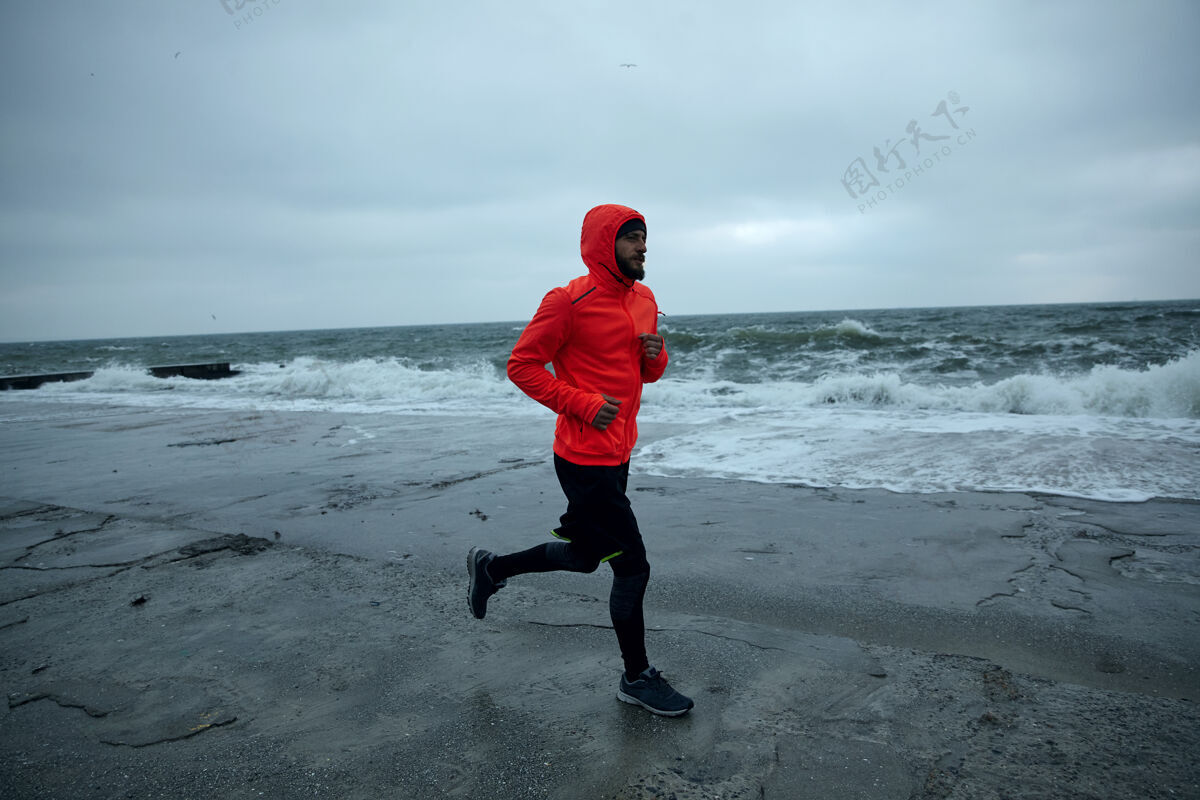 跑步一个留着浓密胡须的年轻人在寒冷的清晨在沙滩上训练 慢跑时穿着黑色运动服和带兜帽的暖橙色外套健身和运动理念健康阴天海滩