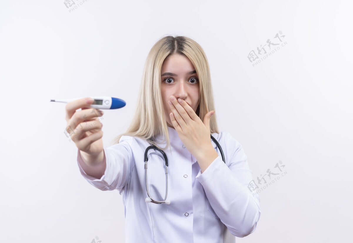抱着惊讶的医生年轻的金发女孩戴着听诊器和医用长袍 拿着温度计对着被摄像机遮住的嘴 在孤立的白色背景下听诊器惊讶嘴巴