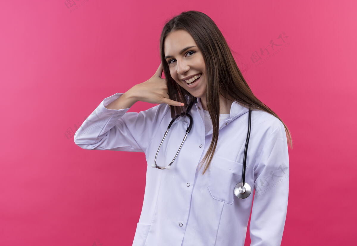 听诊器微笑的年轻医生女孩穿着听诊器医用长袍 在孤立的粉红色背景上显示打电话的手势年轻女孩粉色