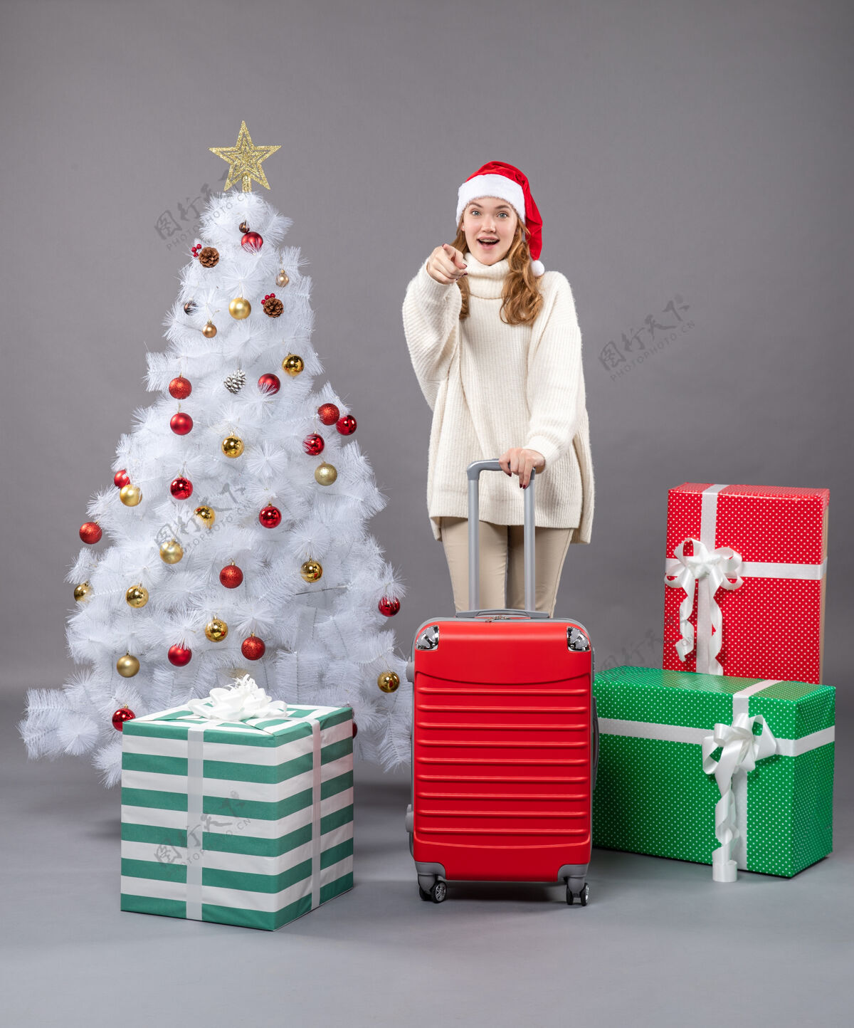 圣诞帽正面图：站在圣诞树旁的快乐的小女孩 带着红色的手提箱礼物站立庆祝